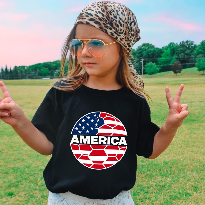 May include: A black short-sleeved t-shirt featuring a circular graphic on the chest. The graphic displays a stylized soccer ball pattern with the top half in blue with white stars and the bottom half in red and white stripes, with the word AMERICA.