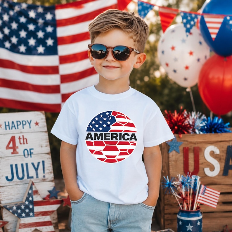 May include: A white short-sleeved t-shirt featuring a circular graphic on the chest. The graphic displays a stylized American flag pattern in red, white, and blue with the word AMERICA printed in bold black capital letters across the center.