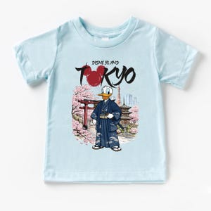 May include: Light blue t-shirt featuring a graphic of Donald Duck in a traditional Japanese kimono, with the text "Disneyland Tokyo". The design includes cherry blossoms, a pagoda, and a red torii gate.
