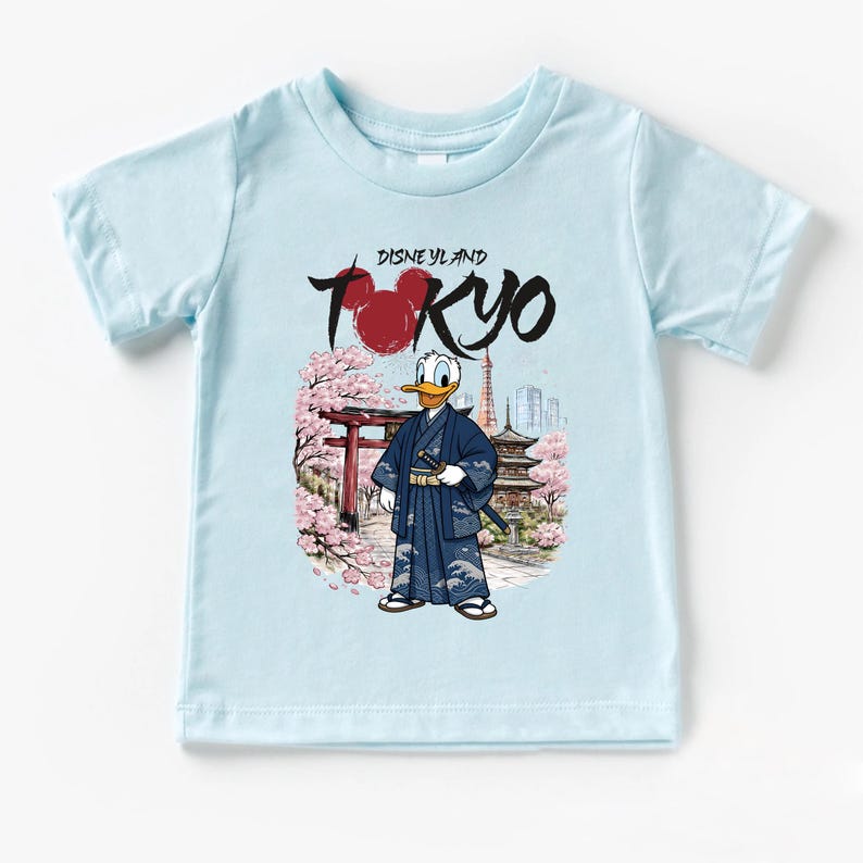 May include: Light blue t-shirt featuring a graphic of Donald Duck in a traditional Japanese kimono, with the text "Disneyland Tokyo". The design includes cherry blossoms, a pagoda, and a red torii gate.