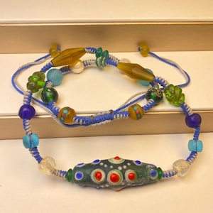 Peut inclure: Deux bracelets de perles faits à la main. L'un comporte une perle oblongue vert foncé avec des accents rouges et bleus. L'autre a un cordon bleu et gris avec diverses perles colorées, dont vertes, jaunes et bleues. Les bracelets sont exposés sur une surface blanche.