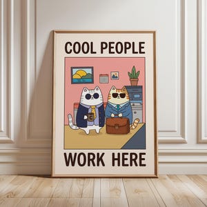 May include: Framed print featuring two cartoon cats in business attire, wearing sunglasses. The image has the text "COOL PEOPLE WORK HERE" in bold letters. The background is a pink office setting with a desk, a small painting, and a potted plant.