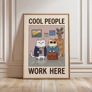 Puede incluir: Un póster enmarcado con dos gatos de dibujos animados vestidos con trajes. Un gato sostiene una taza de café, el otro lleva gafas de sol y un maletín. El texto "COOL PEOPLE WORK HERE" se muestra en la parte superior e inferior de la imagen.
