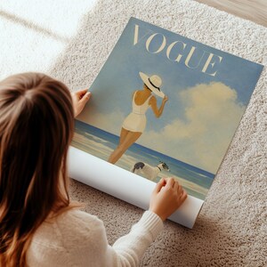 May include: A rolled-up Vogue poster featuring a vintage illustration of a woman in a white swimsuit and hat on a beach with two dogs. The poster has a blue and beige color scheme.