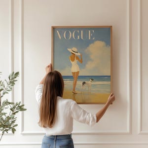 May include: A framed print of a vintage Vogue magazine cover. The artwork depicts a woman in a white swimsuit and hat on a beach with a dog. The word "VOGUE" is at the top in a light blue sky.