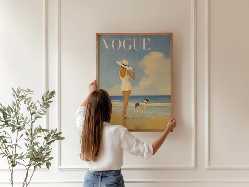 May include: A framed print of a vintage Vogue magazine cover. The artwork depicts a woman in a white swimsuit and hat on a beach with a dog. The word "VOGUE" is at the top in a light blue sky.