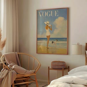 May include: A framed Vogue magazine cover art print. The artwork depicts a person in a white swimsuit and hat standing on a beach with a dog. The word "VOGUE" is at the top in white letters. The background features a blue sky and ocean.