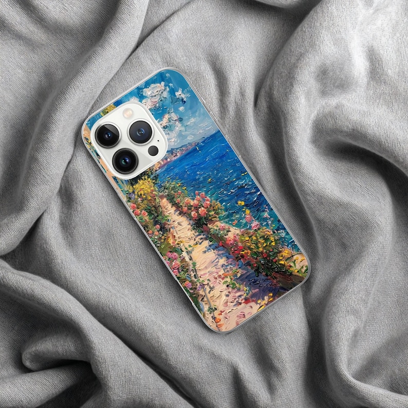 May include: A phone case featuring a vibrant impressionist painting of a coastal scene. The artwork depicts a path lined with colorful flowers overlooking a bright blue sea under a cloudy sky. The case is made of clear plastic.