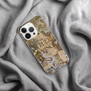 May include: A clear phone case featuring a collage design with the text "The Cruel Prince." The design includes an illustration of a snake, an owl, and other fantasy elements. The case is on a gray fabric surface.