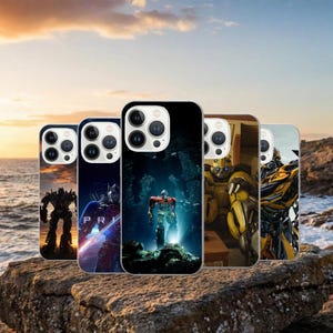 Op de afbeelding: Een verzameling telefoonhoesjes met verschillende Transformers-personages. De hoesjes tonen afbeeldingen van Optimus Prime, Bumblebee en andere Autobots en Decepticons. De hoesjes worden getoond op een rotsachtig oppervlak met een zonsondergang op de achtergrond.