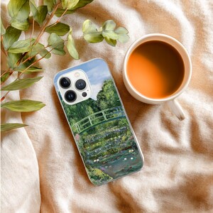 May include: A phone case featuring a painting of a green bridge over a pond with water lilies, trees, and a blue sky. A white mug of coffee sits next to the phone case on a beige surface, with green leaves in the upper left corner.