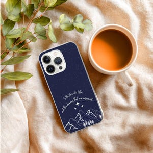 May include: A navy blue phone case with a starry sky design and white mountain and tree illustrations. The case has the quote, "To the stars who listen and the dreams that are answered." A cup of coffee sits nearby.