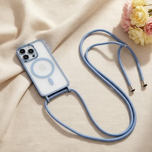 以下が含まれることがあります： ライトブルーの縁取りと、お揃いのブルーのストラップが付いた透明な携帯電話ケース。ケースの背面には円形のデザインがあります。ストラップにはシルバーカラーの金属チップが付いています。