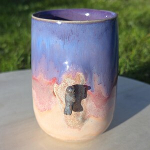 Puede incluir: Un vaso de cerámica hecho a mano con un grifo único. El vaso presenta un degradado de colores, incluyendo morado, azul, rojo y crema. El grifo es un pequeño accesorio metálico. El vaso probablemente se utiliza para bebidas.