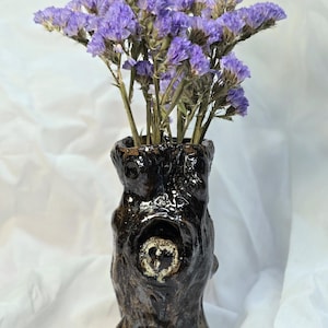 Può includere: Un vaso in ceramica marrone scuro con una superficie ruvida simile alla corteccia e una figura di gufo scolpita all'interno di un'apertura cava. Il vaso è riempito con un bouquet di fiori di statice viola, creando un contrasto tra vaso e fiori.