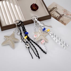 May include: Two keychains featuring a starfish charm with gold studs, a silver-toned key, and colorful beads attached to a braided cord. One keychain has a black cord with white accents, while the other has a white cord with black accents.