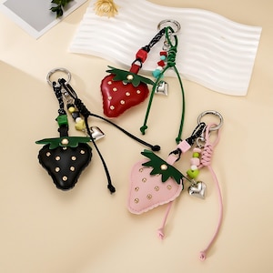 May include: Three strawberry-shaped keychains in red, black, and light pink. Each features a green leaf top, gold-tone studs, a silver metal ring, a dangling silver heart charm, and colorful beads attached to a cord with a knotted design.