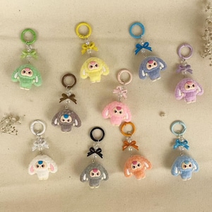Può includere: Una collezione di portachiavi in peluche colorati in varie tonalità pastello, tra cui verde, giallo, rosa, viola, blu, bianco, grigio e arancione. Ogni portachiavi presenta un personaggio dei cartoni animati con un fiocco e un accento di perline.