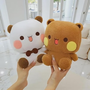 May include: Two plush bear toys. One is white with brown accents and a brown bow tie. The other is brown with yellow cheek accents. Both have smiling faces with pink cheeks and open mouths.