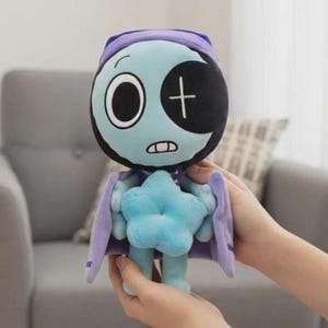 May include: A plush doll with a light blue face, a black eye patch, and a crescent moon-shaped head covering. The doll has a star-shaped body and wears a purple cape. The doll is held in front of a gray couch.