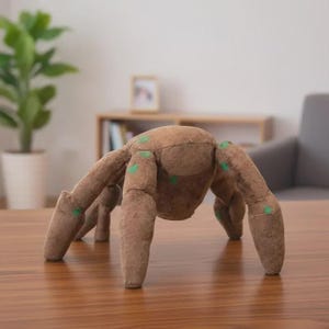 May include: A brown plush spider toy with eight legs and green spots. The toy is made of soft material and is sitting on a wooden table. The spider is a whimsical decorative item.