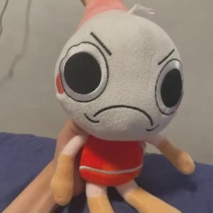 Puede incluir: Un peluche con cabeza blanca, ojos negros grandes y expresión de ceño fruncido. Tiene un torso rojo, brazos y piernas color canela, y una aleta rosa y canela en la parte superior. El juguete se sostiene contra una superficie azul.