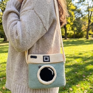 May include: A handmade crochet camera bag in shades of light blue, cream, and black. The bag features a large circular lens and a rectangular top detail, with a cream-colored strap. The bag is worn over the shoulder.