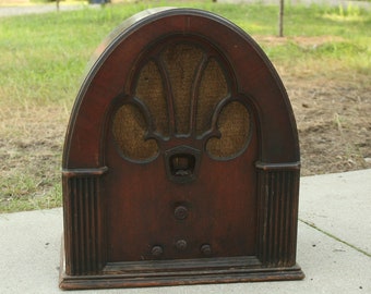 May include: An antique wooden radio with a dark brown finish. The radio has an arched top and a speaker grille with a decorative pattern. The radio is sitting on a concrete surface.