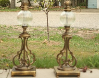 May include: A pair of matching table lamps featuring antique brass-finished metal bases with ornate scrollwork, clear ribbed glass globes, and slender stems topped with white light bulbs. The lamps stand on square bases with small feet.
