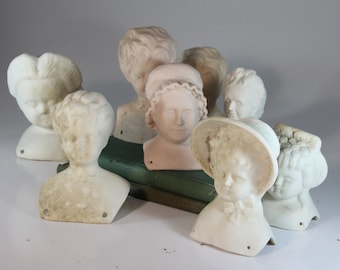 May include: A collection of antique bisque doll heads, featuring various hairstyles and bonnets. The heads are primarily off-white with some showing signs of age and wear. They are arranged on a green surface.