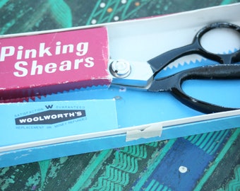 Könnte beinhalten: Eine vintage-Pinkierschere in einer blauen und weißen Schachtel mit dem Text "Pinking Shears" und "Woolworth's" auf der Schachtel. Die Schachtel trägt außerdem den Hinweis "Satisfaction Guaranteed, Replacement or Money Refunded."