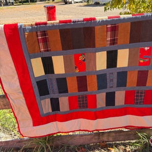 May include: A handmade quilt with a patchwork design, featuring a variety of fabrics in shades of brown, red, and black. The quilt has a red border and is hanging on a wooden fence.