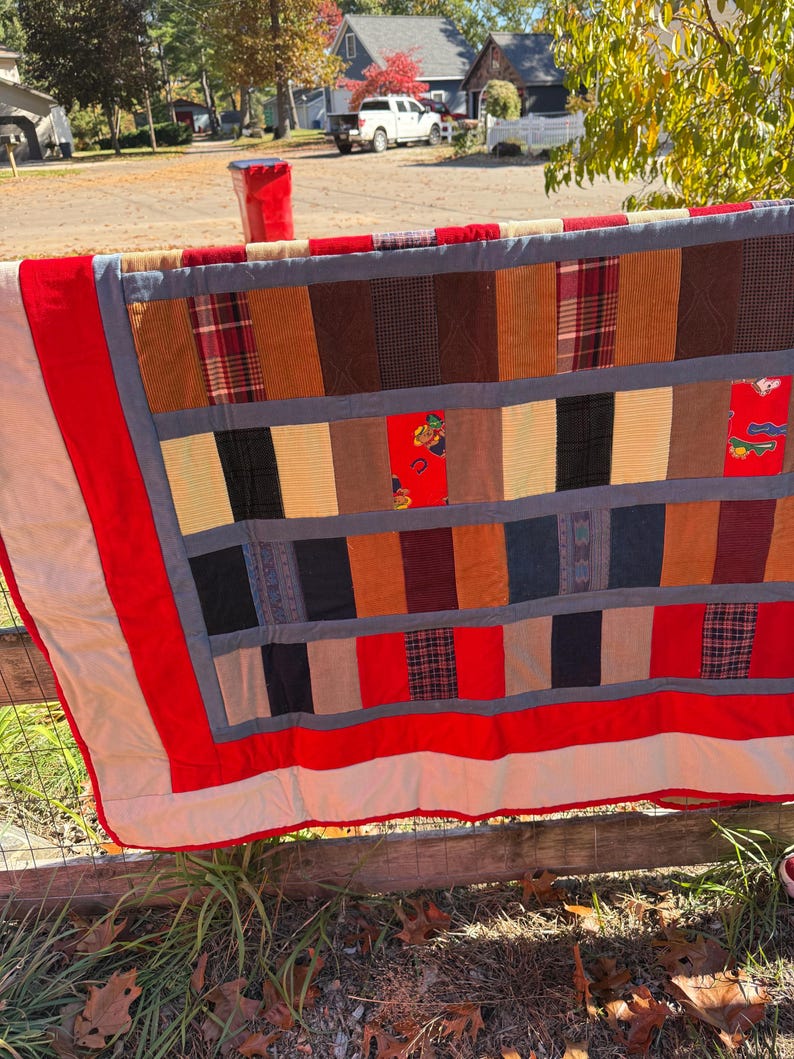 May include: A handmade quilt with a patchwork design, featuring a variety of fabrics in shades of brown, red, and black. The quilt has a red border and is hanging on a wooden fence.