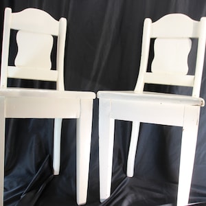 May include: Two white wooden chairs with a classic design. Each chair has a rectangular seat, four legs, and a backrest with a decorative cutout. The chairs are painted white and appear to be vintage or antique.