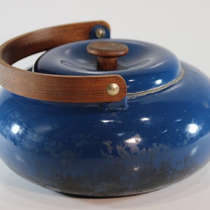 May include: A blue enamel teapot with a wooden handle and lid knob. The handle is a curved, light brown wood attached with brass rivets. The teapot has a rounded shape and a shiny finish.