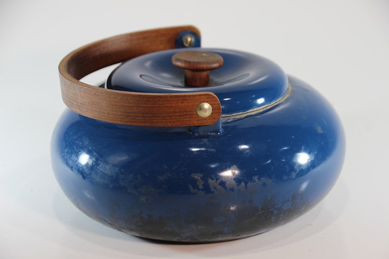 May include: A blue enamel teapot with a wooden handle and lid knob. The handle is a curved, light brown wood attached with brass rivets. The teapot has a rounded shape and a shiny finish.