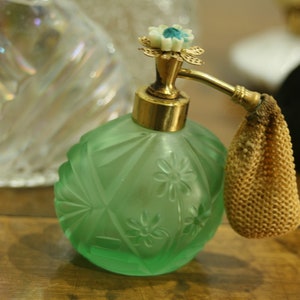 May include: A vintage green glass perfume bottle with a floral design and gold accents. The bottle has a decorative sprayer and a small, beige mesh pouch. Other glass bottles are in the background.