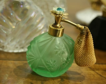Könnte beinhalten: Eine Vintage-Parfümflasche aus grünem Glas mit floralem Design und goldenen Akzenten. Die Flasche hat einen dekorativen Zerstäuber und einen kleinen, beigefarbenen Netzbeutel. Weitere Glasflaschen sind im Hintergrund.