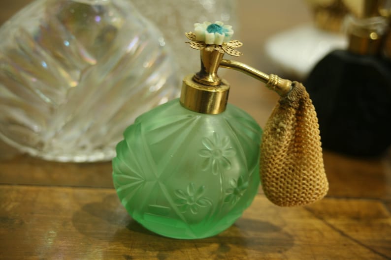 May include: A vintage green glass perfume bottle with a floral design and gold accents. The bottle has a decorative sprayer and a small, beige mesh pouch. Other glass bottles are in the background.