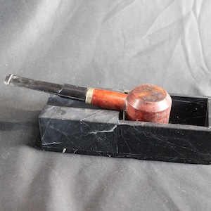 Puede incluir: Una pipa de tabaco con un cuenco marrón oscuro y una boquilla negra, apoyada en un cenicero de mármol negro. La pipa tiene un cuenco de madera marrón rojizo y una banda dorada. El cenicero es rectangular.
