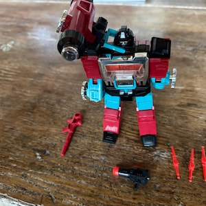 Op de afbeelding: Een rode, zwarte en blauwgroene robotactiefiguur met accessoires. De robot heeft een groot kanon op zijn schouder en verschillende kleine rode wapens. De figuur staat op een houten oppervlak.