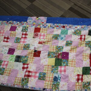 May include: A colourful patchwork quilt featuring squares of various patterns and hues, including pink, red, green, and plaid. The quilt is pinned to a blue fabric border. A handmade textile.