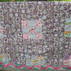 May include: A handmade quilt with a patchwork design. The quilt features a mix of patterns, including floral, geometric, and abstract shapes in shades of pink, brown, and white. The quilt is displayed outdoors, draped over a wooden fence.