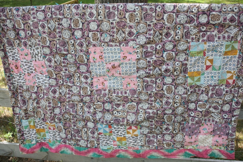 May include: A handmade quilt with a patchwork design. The quilt features a mix of patterns, including floral, geometric, and abstract shapes in shades of pink, brown, and white. The quilt is displayed outdoors, draped over a wooden fence.