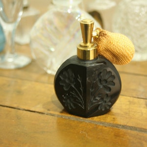 May include: Black perfume bottle with a gold-colored atomizer and a decorative, textured bulb. The bottle has a floral design carved into the surface. Other glass bottles are in the background.