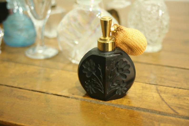 May include: Black perfume bottle with a gold-colored atomizer and a decorative, textured bulb. The bottle has a floral design carved into the surface. Other glass bottles are in the background.
