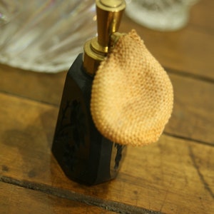 May include: A vintage black perfume bottle with a gold-colored top and a beige powder puff. The bottle has a decorative floral design and sits on a wooden surface. The perfume bottle is an antique item.