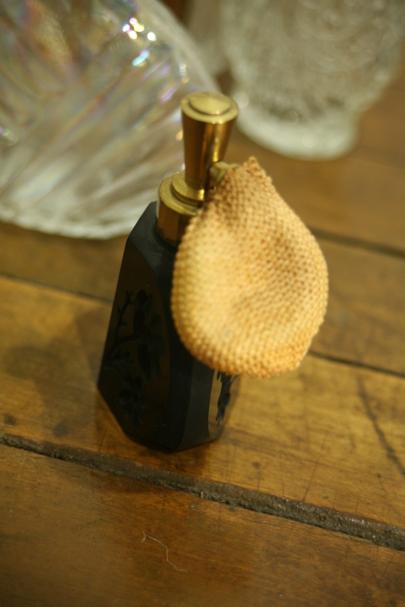 May include: A vintage black perfume bottle with a gold-colored top and a beige powder puff. The bottle has a decorative floral design and sits on a wooden surface. The perfume bottle is an antique item.