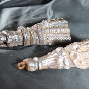 Könnte beinhalten: Zwei antike architektonische Elemente aus Gusseisen mit einer verwitterten, dunkelbraunen Oberfläche. Jedes Stück hat ein detailliertes, strukturiertes Design mit seilartigen und rechteckigen Mustern. Die Elemente liegen auf einer dunklen, strukturierten Oberfläche.