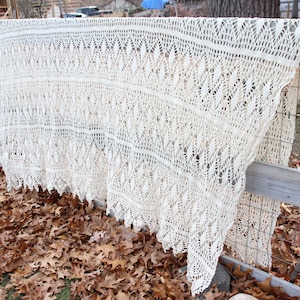 May include: A cream-colored, open-weave shawl with intricate lace detailing is draped over a wooden fence. The shawl's design features repeating patterns and a scalloped edge. The background shows fallen leaves.